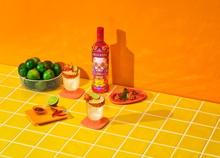 "Una botella de Smirnoff Spicy Tamarind junto a un cóctel, un plato de comida picante y un tazón de limones sobre un fondo naranja.
