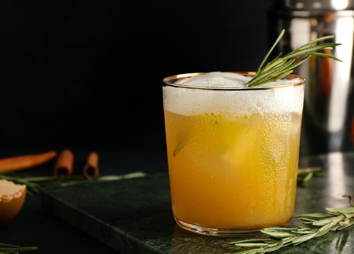 Cóctel amarillo sobre la barra de trabajo con espuma y guarnición de hierbas, con herramientas de bar en el fondo.