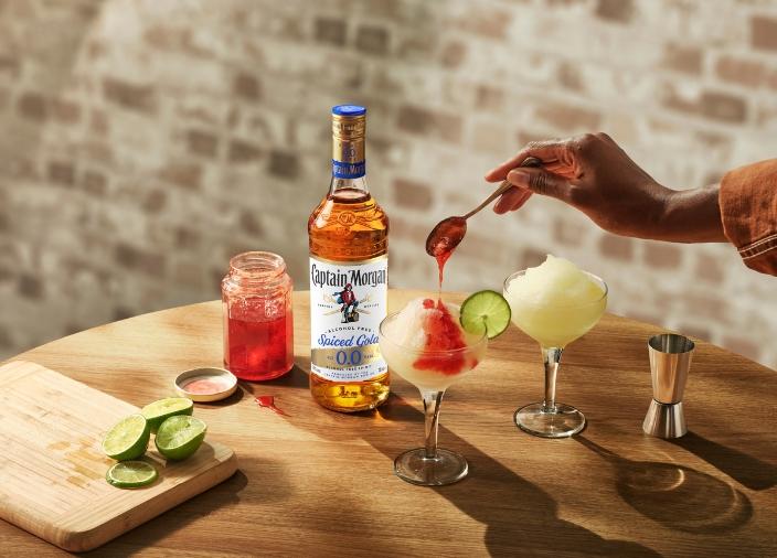 Botella de Captain Morgan Spiced Gold 0,0% sin alcohol sobre una mesa rodeada de alimentos y bebidas variadas sobre un fondo rojo.