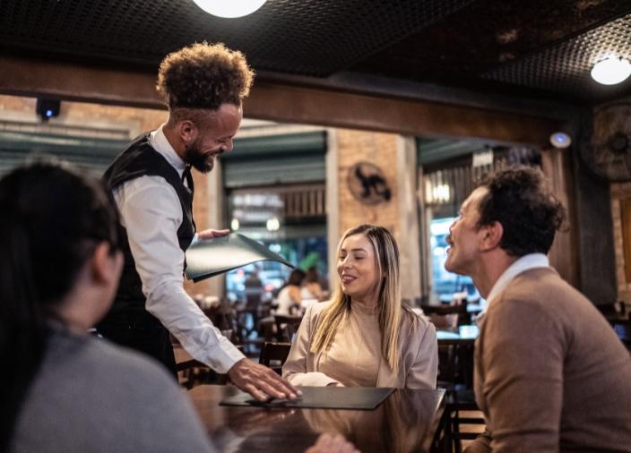 Un camarero sonriente atendiendo a una invitada que está sentada en una mesa con dos amigos mientras recoge el menú frente a ella.