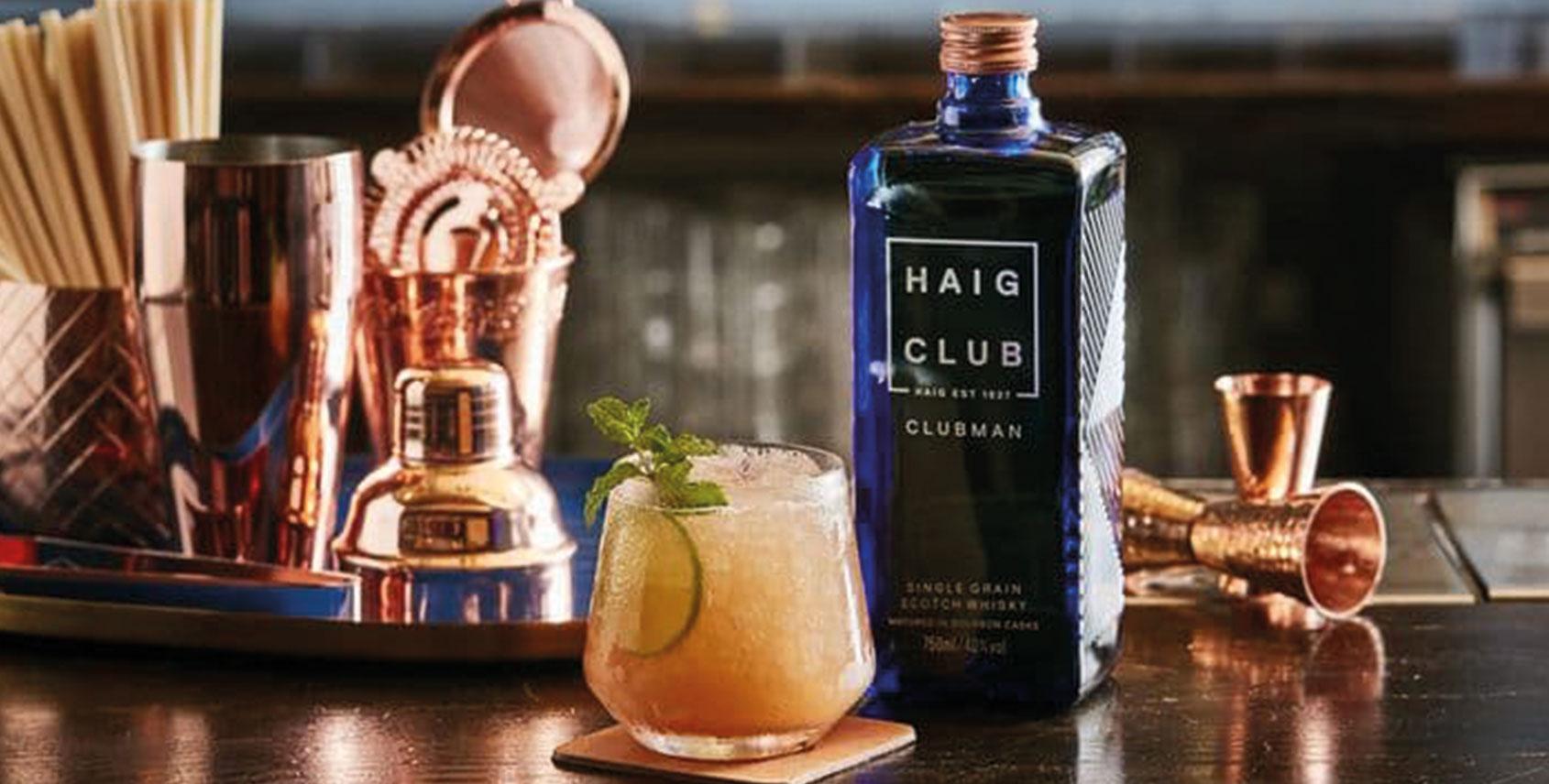 Botella Haig Club al lado un vaso servido y decorado de menta y alrededor utensilios