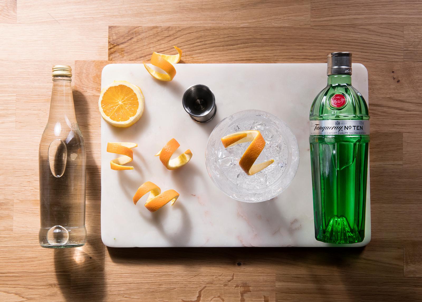 A filled gin goblet with lime garnish next to a bottle of Tanqueray, bottles of tonic, sliced limes and a pile of limes in background.