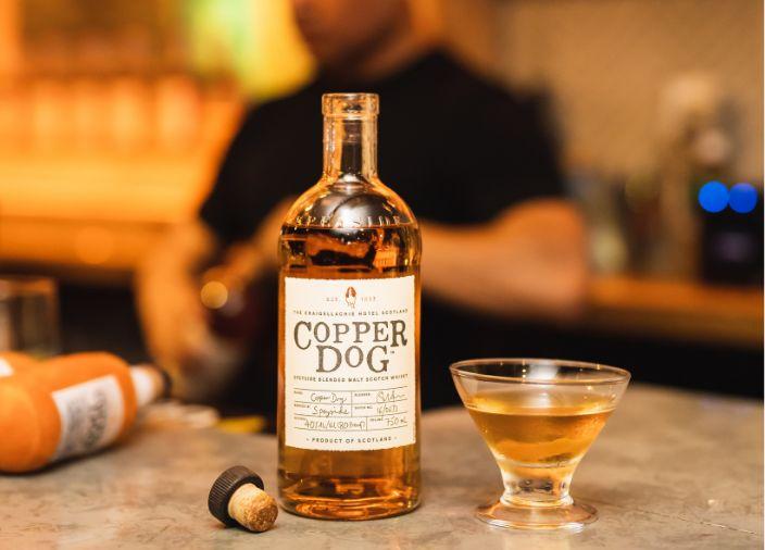 An open bottle of copper dog with a glass of neat whisky, the bartender in the background