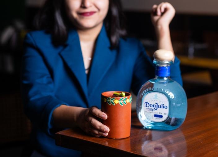 Una mujer sonriendo, sosteniendo una taza de arcilla estampada que contiene un cóctel sobre una mesa, con una botella Don Julio Blanco al lado