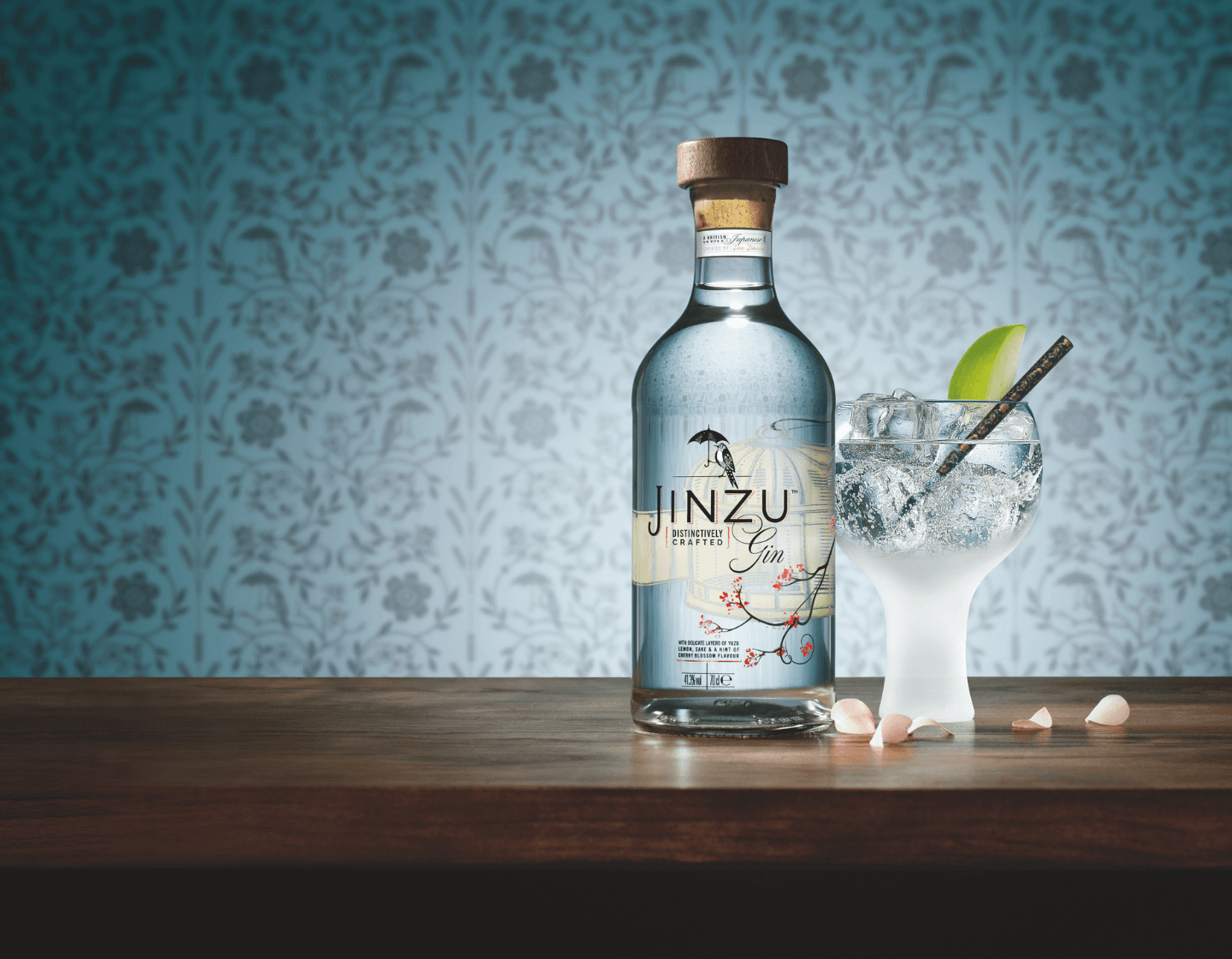 Botella de ginebra Jinzu sobre una mesa junto a un vaso de gin-tonic sobre un fondo de papel pintado con flores azules 