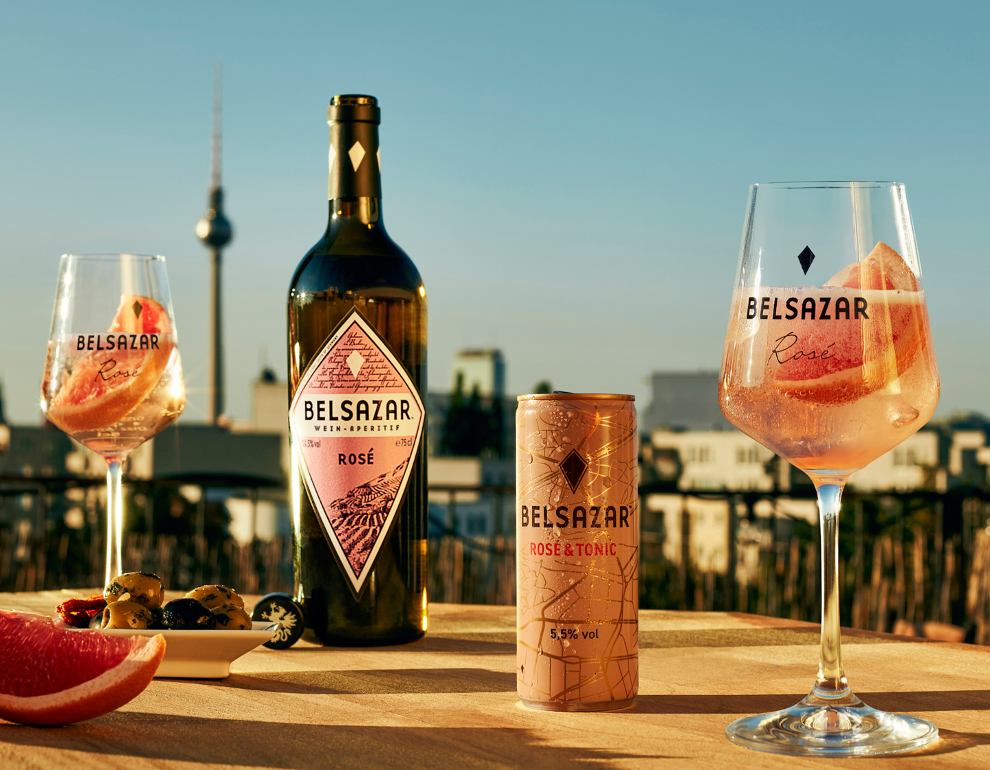 Flasche Belsazar Rosé draußen auf einem Tisch bei Sonnenuntergang neben zwei Weingläsern, die mit rosa Getränken gefüllt sind.