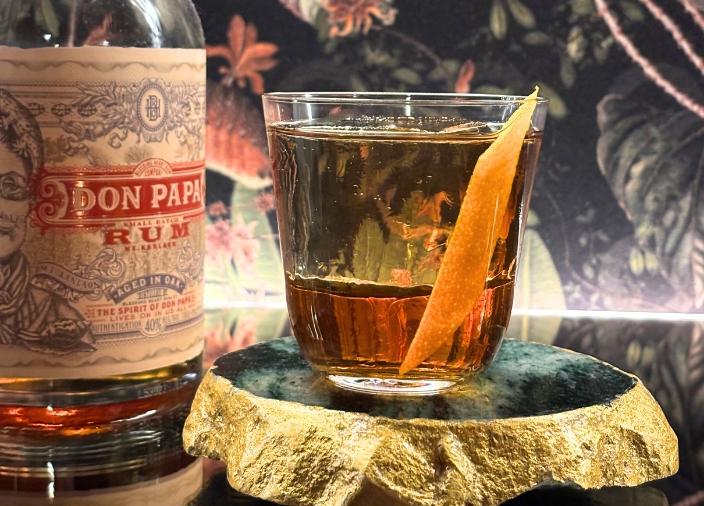 Un cóctel de ron Old Fashioned con una guarnición de piel de naranja dentro de la bebida, junto a una botella de Don Papa Rum.