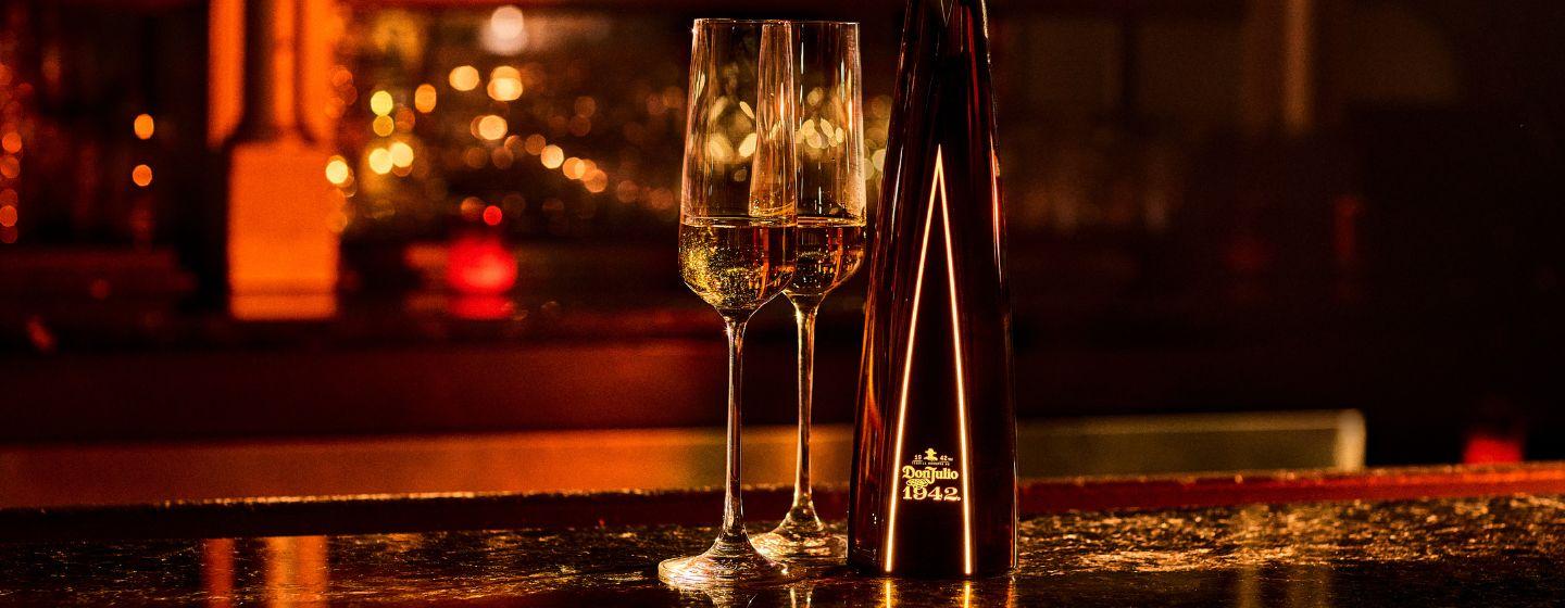 Bottle of Don Julio 1942 tequila against gold curtain beside glass of liquid and ice