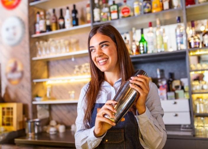 A woman with long dark hair shaking a cocktail shaker behind a bar, smiling.