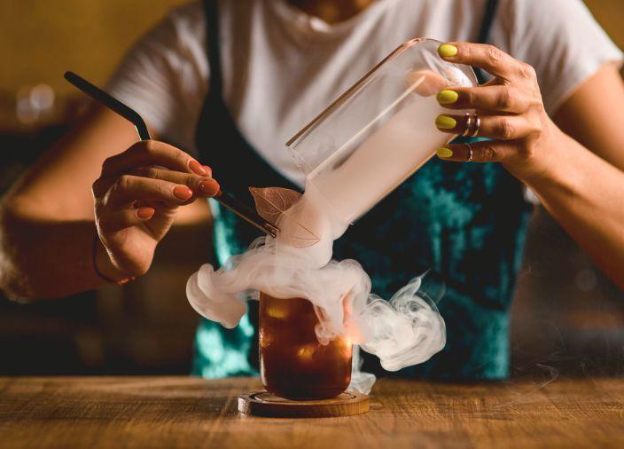 A cocktail glass surrunded by smoke, as bartender arranges garnish.