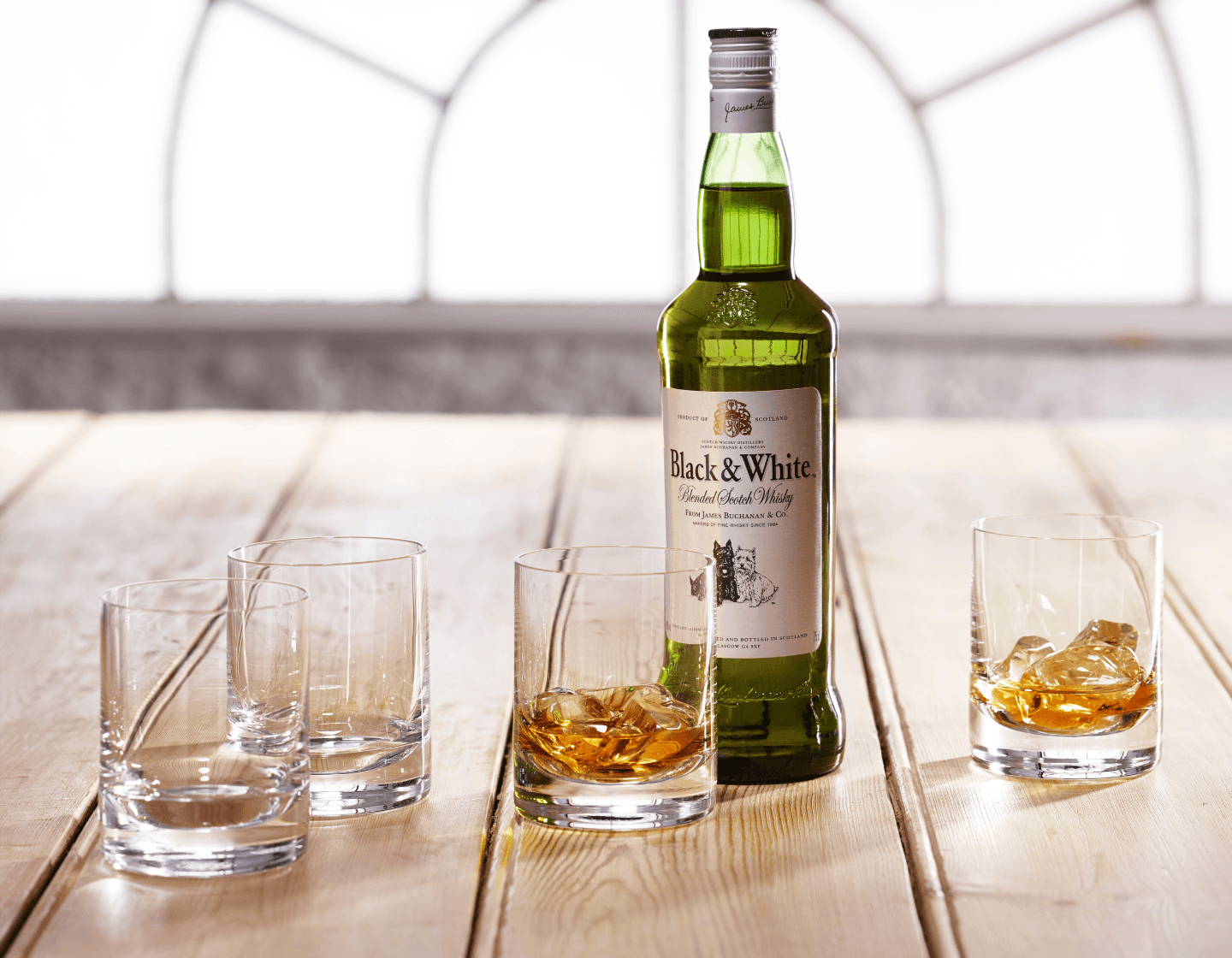 Botella de Black & White sobre una mesa de madera junto a dos vasos de whisky y hielo 