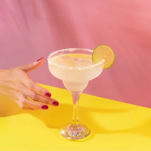 Coquetel margarita com uma fatia de limão como guarnição sendo servido por uma mão feminina sob uma mesa de cor amarela