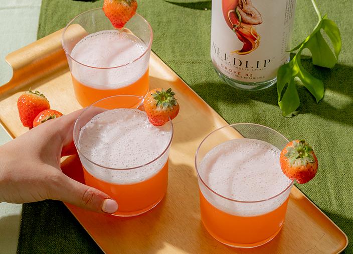 Three glasses of orange cocktail on a wooden board alongside a bottle of Seedlip Grove 42