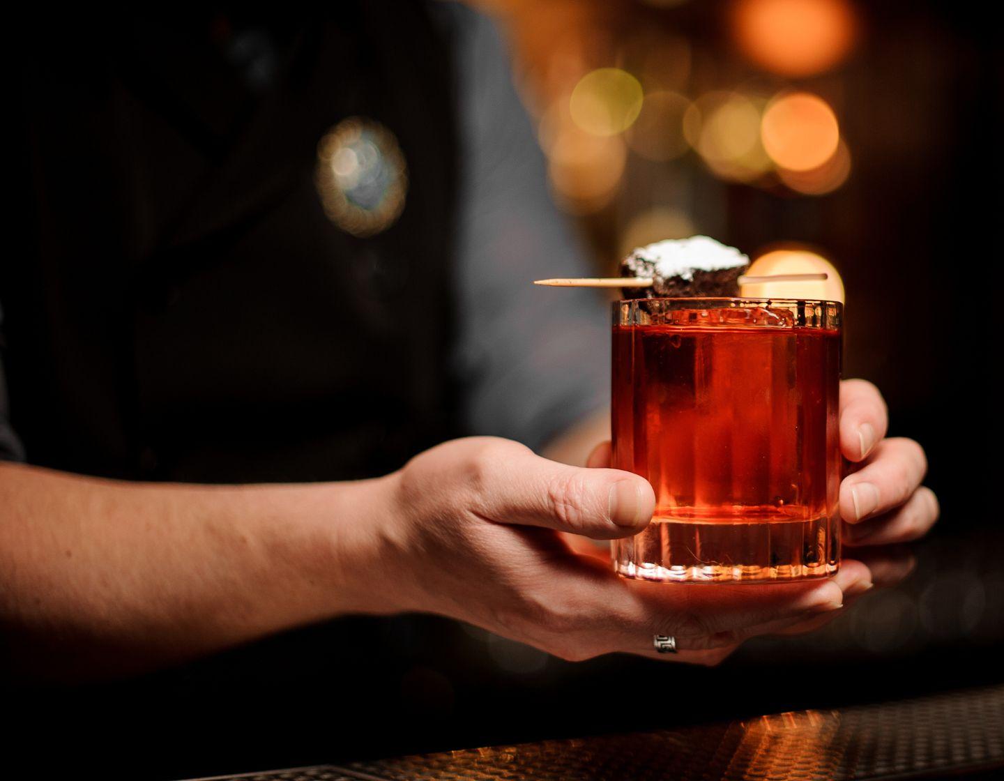 Eine Person hält einen roten Cocktail mit einer Brownie-Garnitur, die mit Puderzucker bestreut ist.