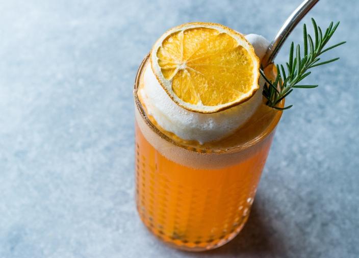 An orange cocktail in a tall glass garnished with a dehydrated orange wheel and a sprig of rosemary.