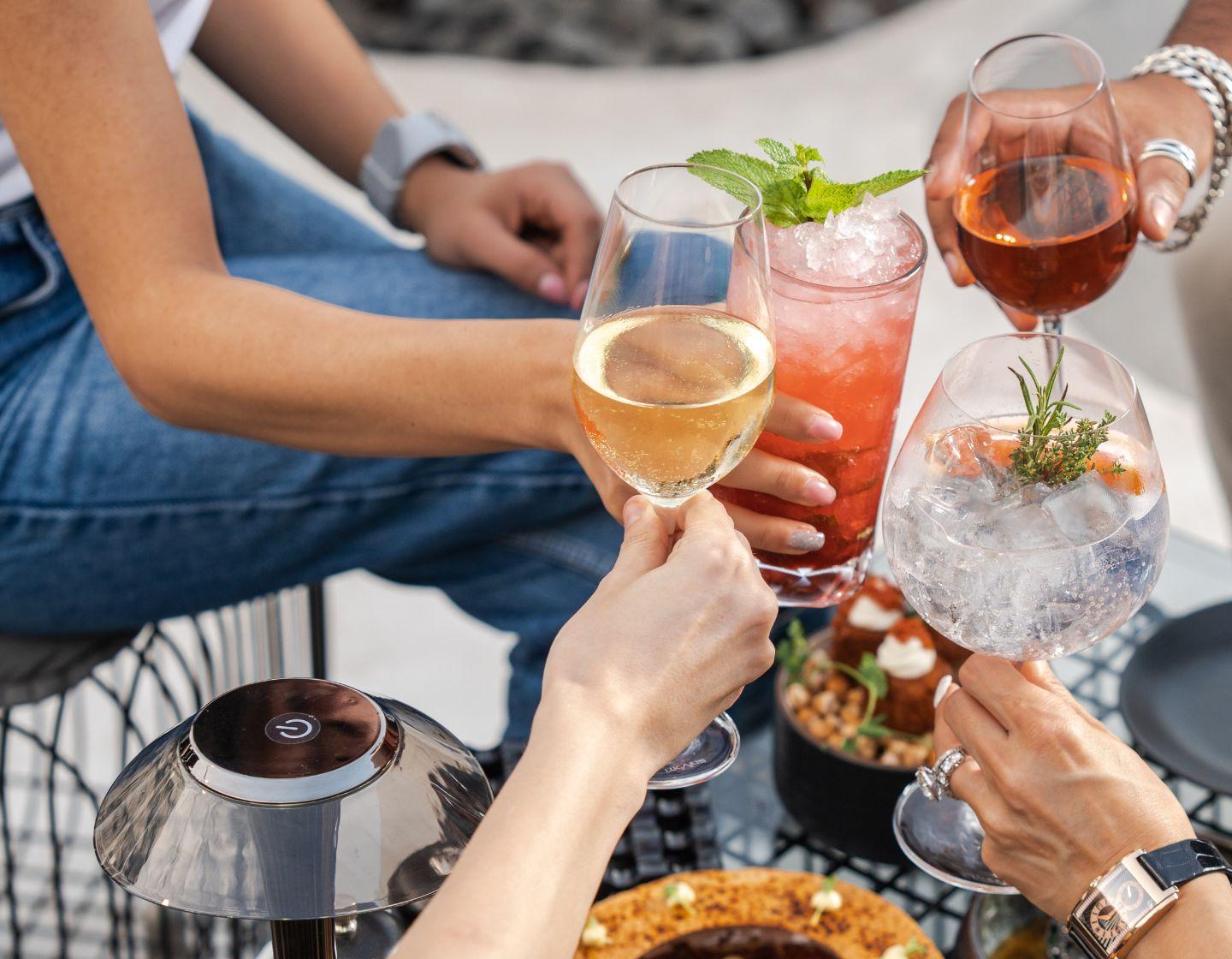 A close-up shot of four different cocktails being clinked together by the arms of four people sitting at a glass table with food.