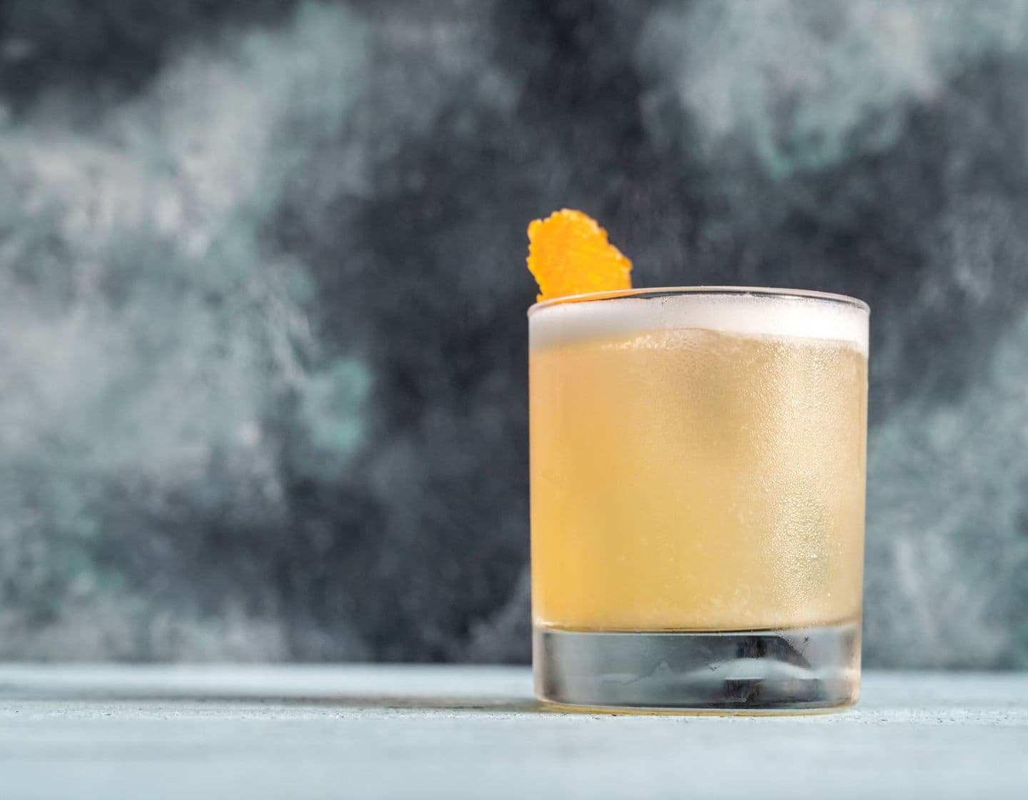 Yellow cocktail with orange garnish on a grey background.