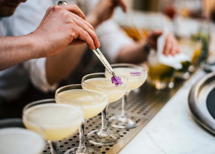 A line of light yellow cocktails in coupette glasses sitting on a bar being garnished.