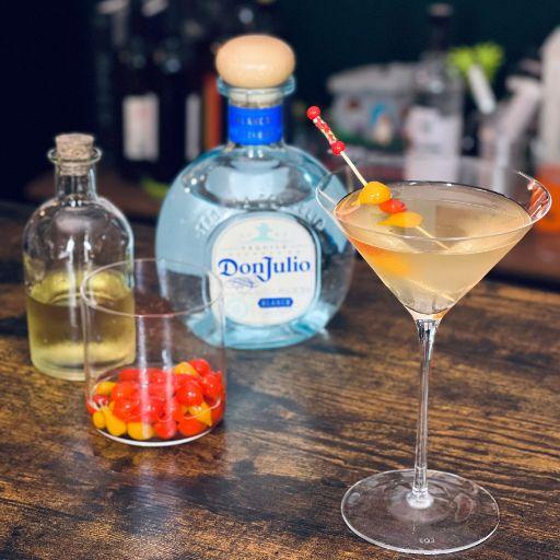 Un cóctel de martini teñido de naranja con guarnición de pepinillos rojos y naranjas y una botella de tequila Don Julio sobre una mesa de madera