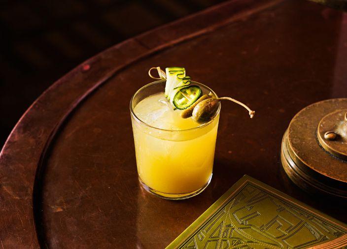 Cóctel amarillo con garnish de alcaparras y pepino.