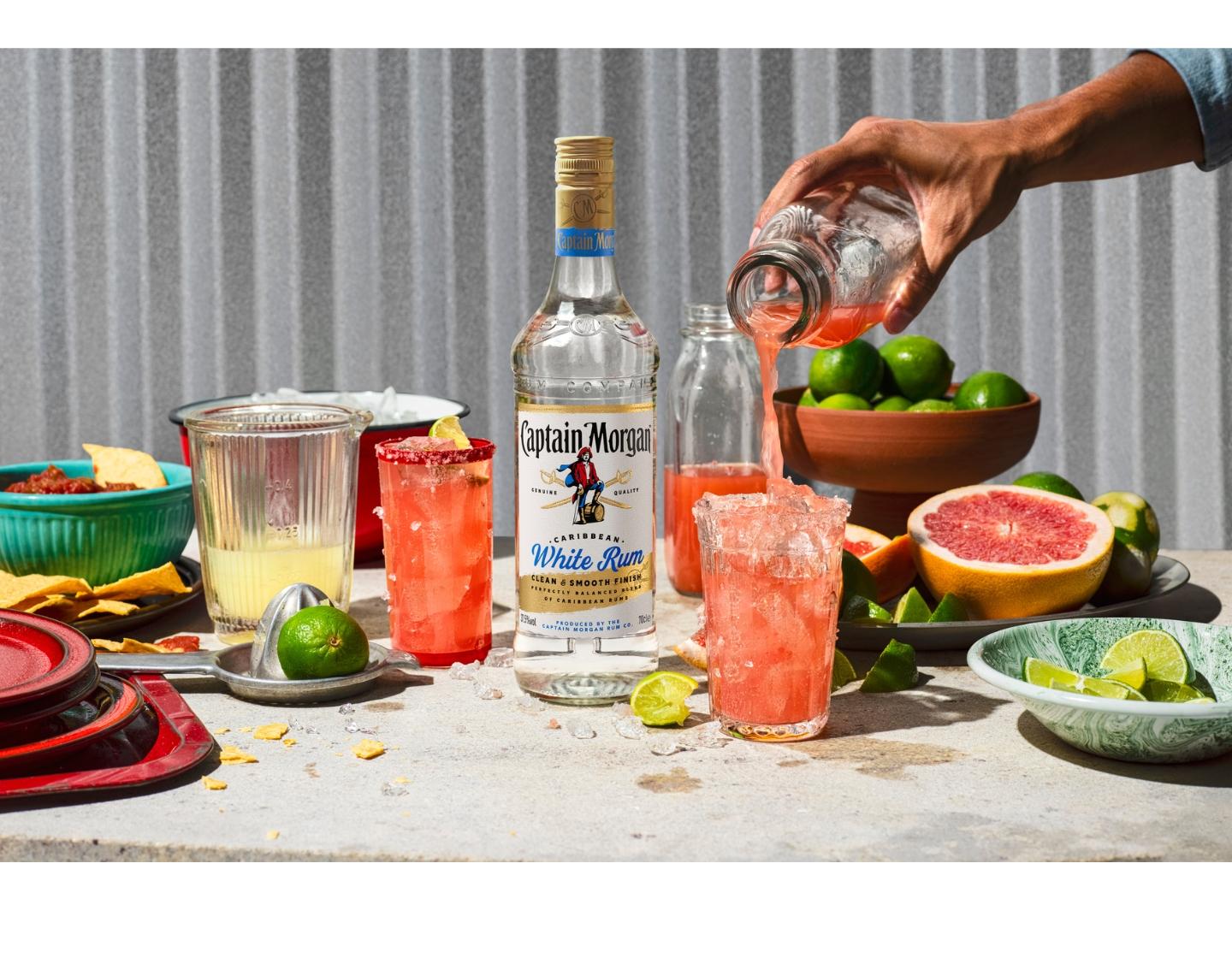 Bottle of Captain Morgan White Rum and two iced glasses of cocktail on a table with a hand filling one of the glasses