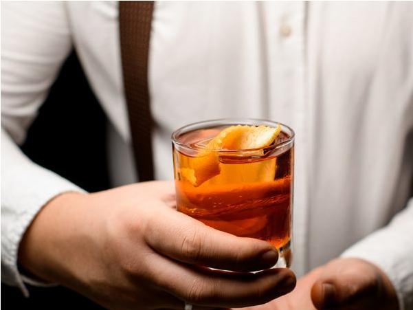 Un bartender con camisa blanca sosteniendo un cóctel dorado con una rodaja de naranja como adorno.