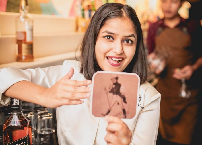 Aashi Bhatnagar sosteniendo un mantel individual Johnnie Walker mientras se apoya en una botella de whisky Black Label Johnnie Walker detrás de la barra