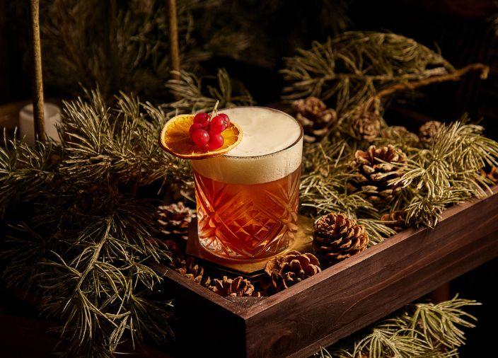 Un cóctel de naranja con espuma blanca y garnish de rodaja de naranja deshidratada.