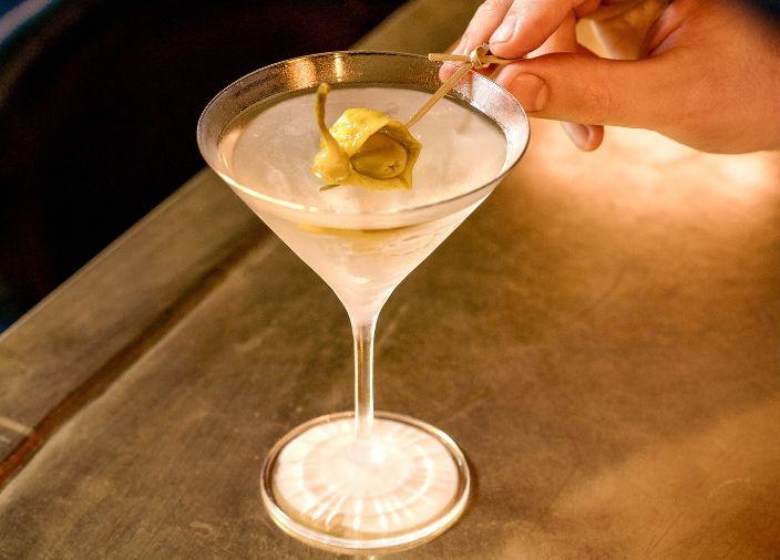 Un martini con la mano de un bartender agregando un garnish de pepinillos verdes.