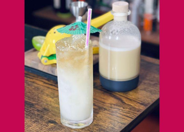 An ombre white and light brown miso cocktail topped with ice, sesame seeds and a green paper umbrella next to a bottle of miso orgeat paste.