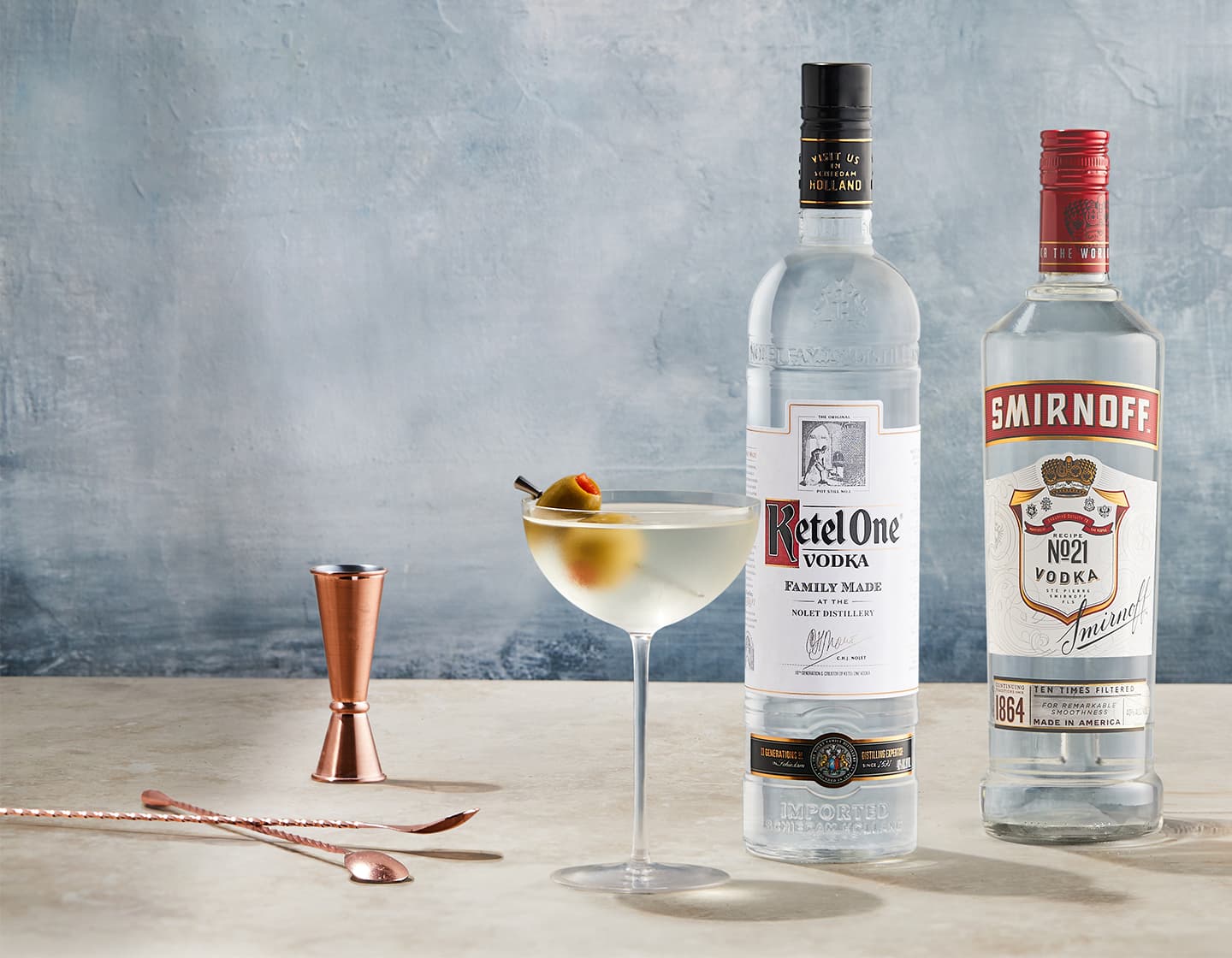 A bottle of Ketel One vodka and Smirnoff vodka on a table, next to a classic cocktail glass, containing a cold drink and a garnish of two olives. A rose gold jigger and stirrer are laying on the tabletop. 