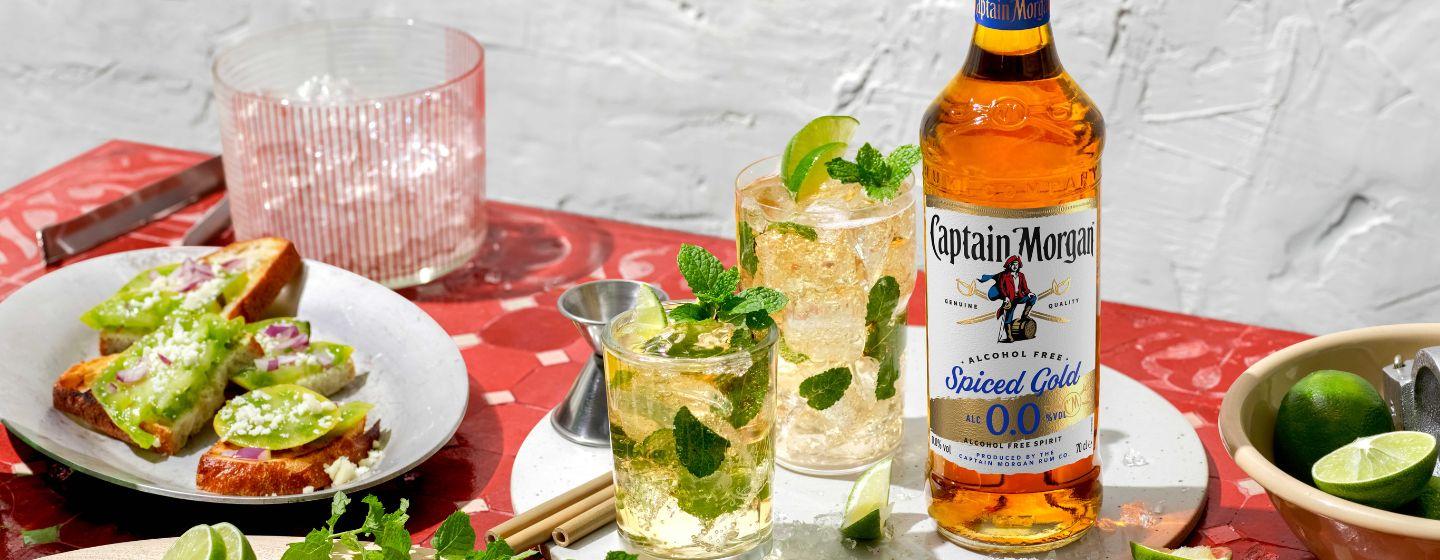 Botella de Captain Morgan Spiced Gold 0.0% Alcohol Free Spirit sobre una mesa rodeada de alimentos y bebidas variadas.