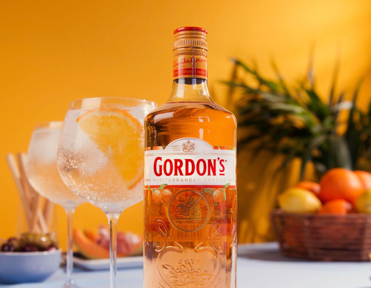  Botella de Gordon's Mediterranean Gin junto a dos copas de G&T sobre un fondo naranja.