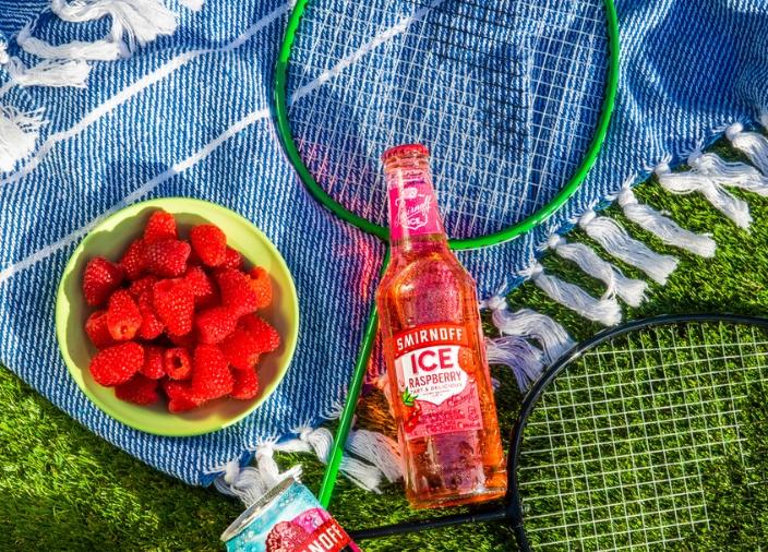 Botella de Smirnoff Ice Raspberry recostada sobre una manta sobre el césped con un plato de frambuesas al lado