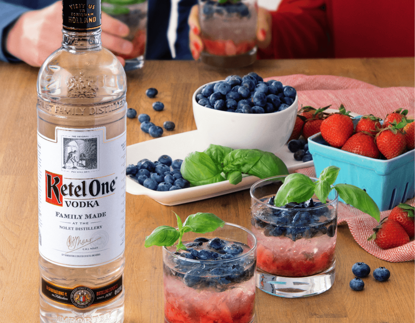 Bottle of Ketel One vodka beside two cocktails filled with blueberries, beside bowls of assorted fruits. 