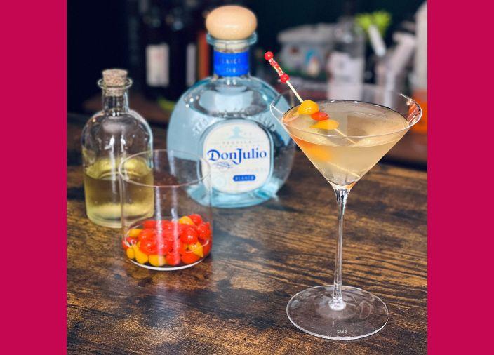 An orange-tinted martini cocktail with a red and orange pickle garnish, and a bottle of Don Julio tequila on a wooden table.