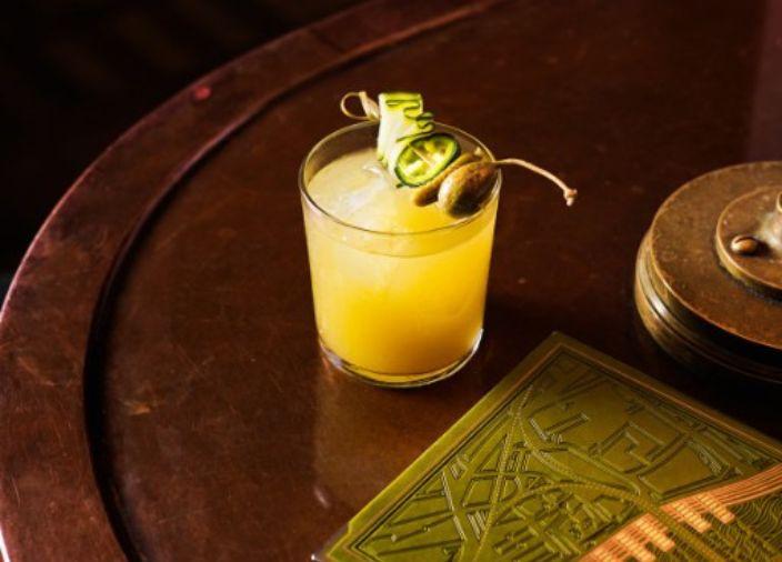 A yellow cocktail with a caperberry and cucumber garnish.