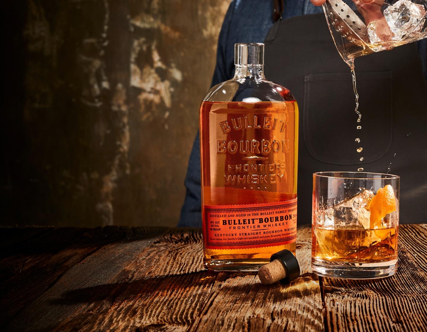Garrafa de Bulleit Bourbon ao lado de um homem servindo o coquetel em um copo 