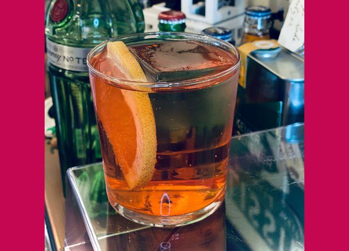 A bright orange cocktail in a short glass with a large format ice cube and a slice of orange.