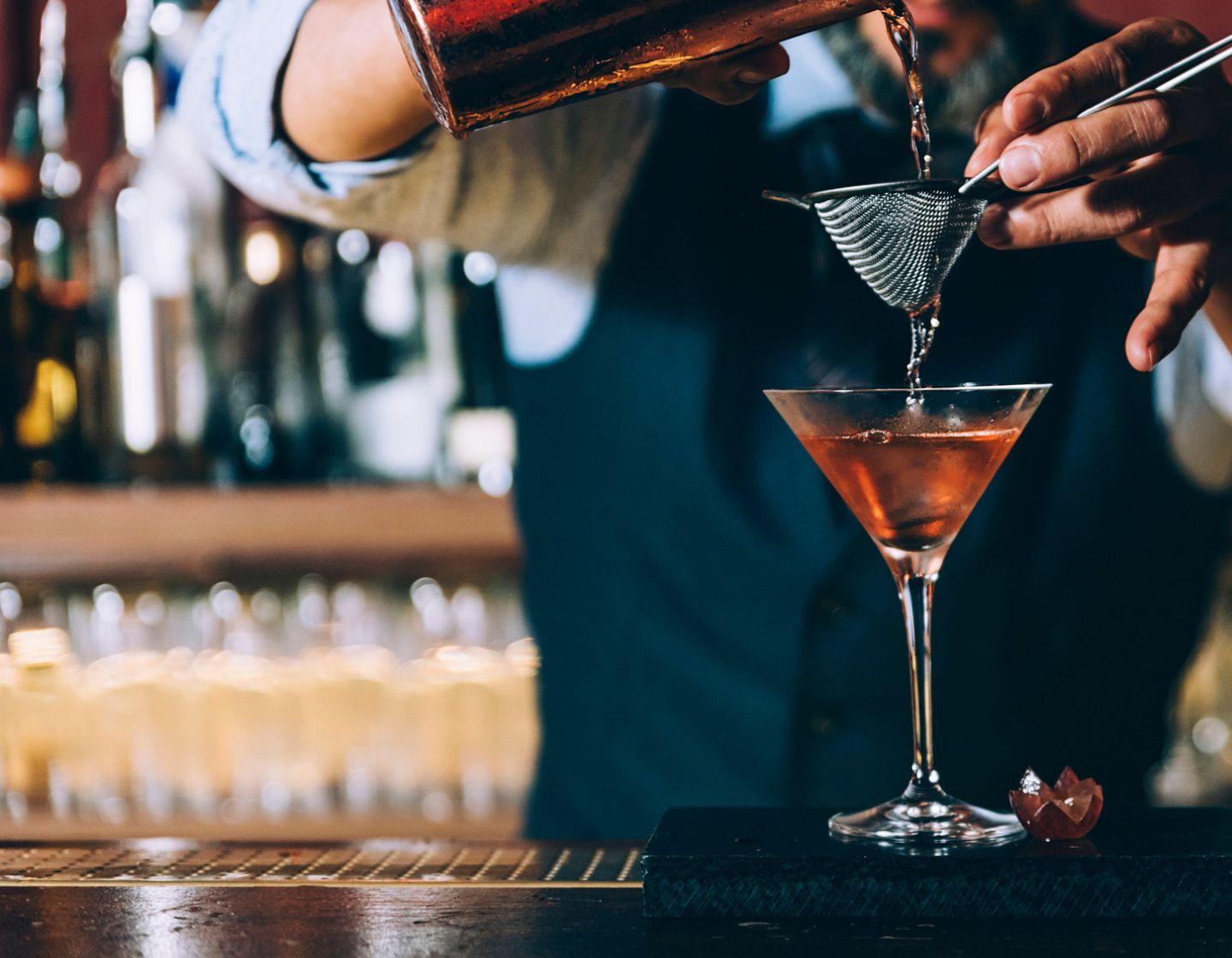 Bartender vertiendo un cóctel dorado desde una coctelera a través de un colador en una copa de cóctel.