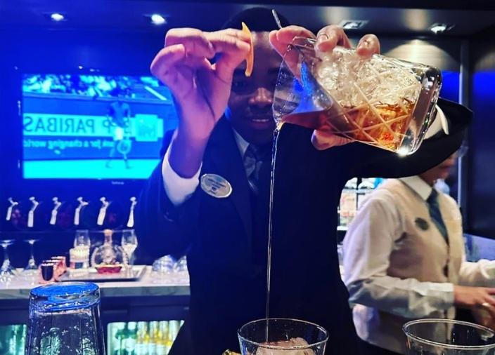 Un hombre detrás de una barra vierte líquido de una jarra con hielo y un colador en un vaso en altura, sosteniendo en la otra mano una guarnición de naranja