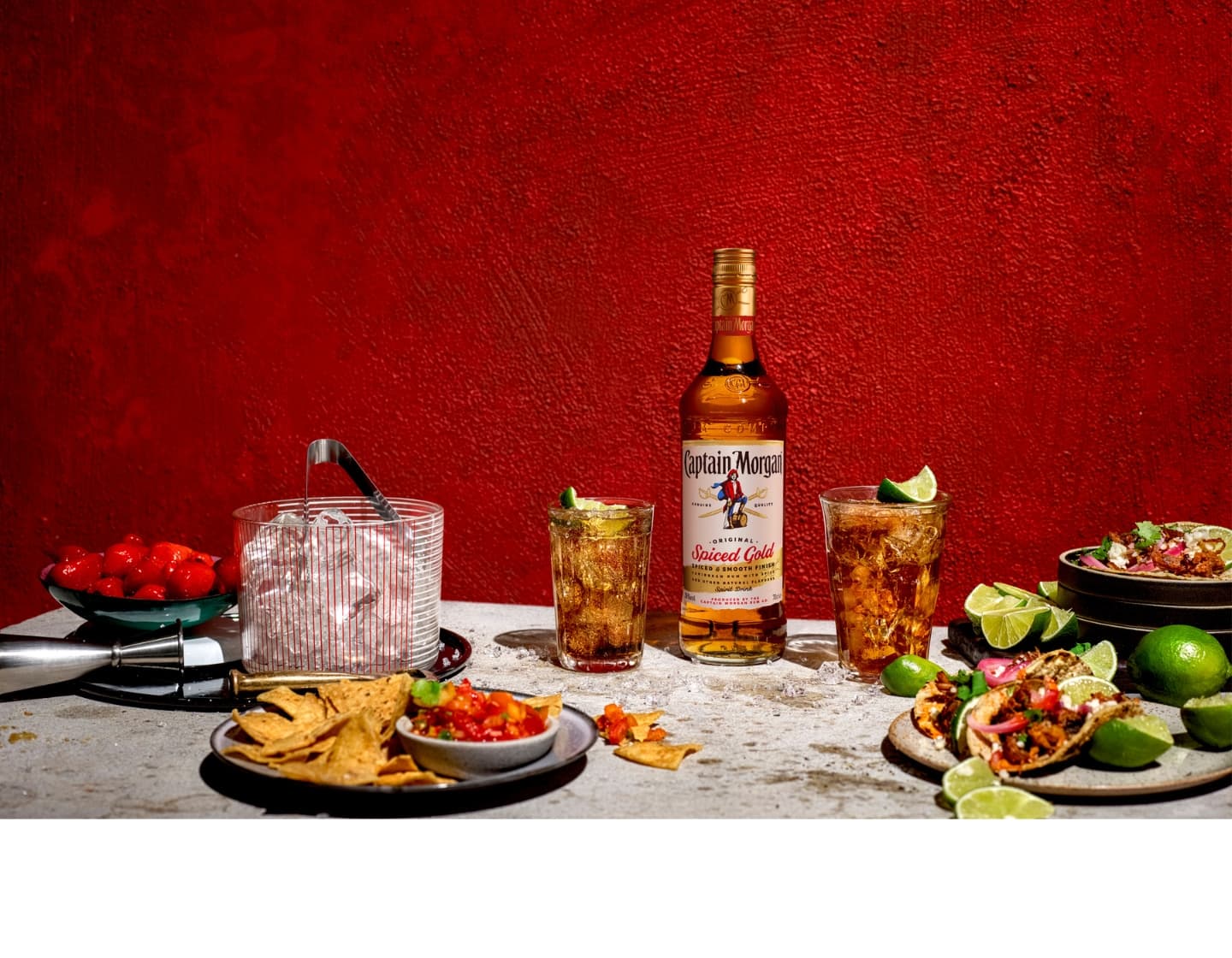 Bottle of Captain Morgan Spiced Gold rum on table beside two iced glasses of rum and coke surrounded by food