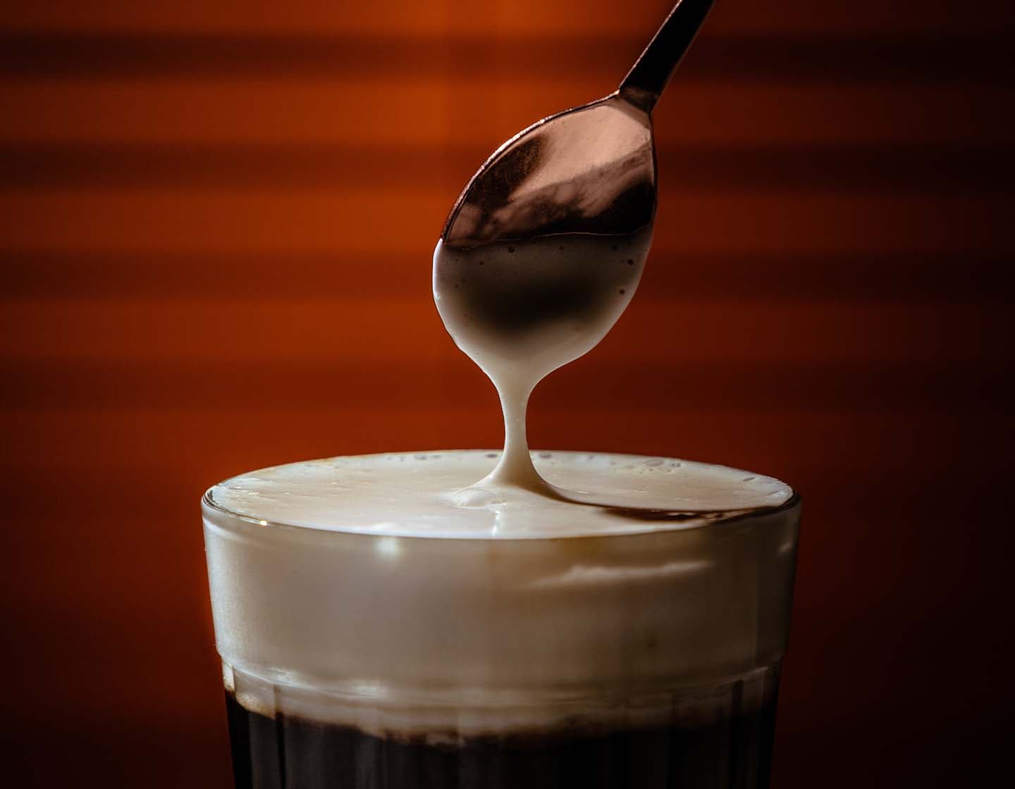 A close-up of a stirring spoon over the top of a drink with a thick layer of cream/foam on top. 