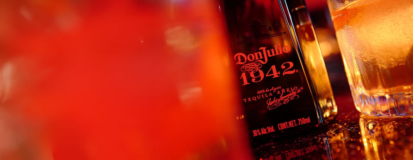 Bottle of Don Julio 1942 tequila against gold curtain beside glass of liquid and ice