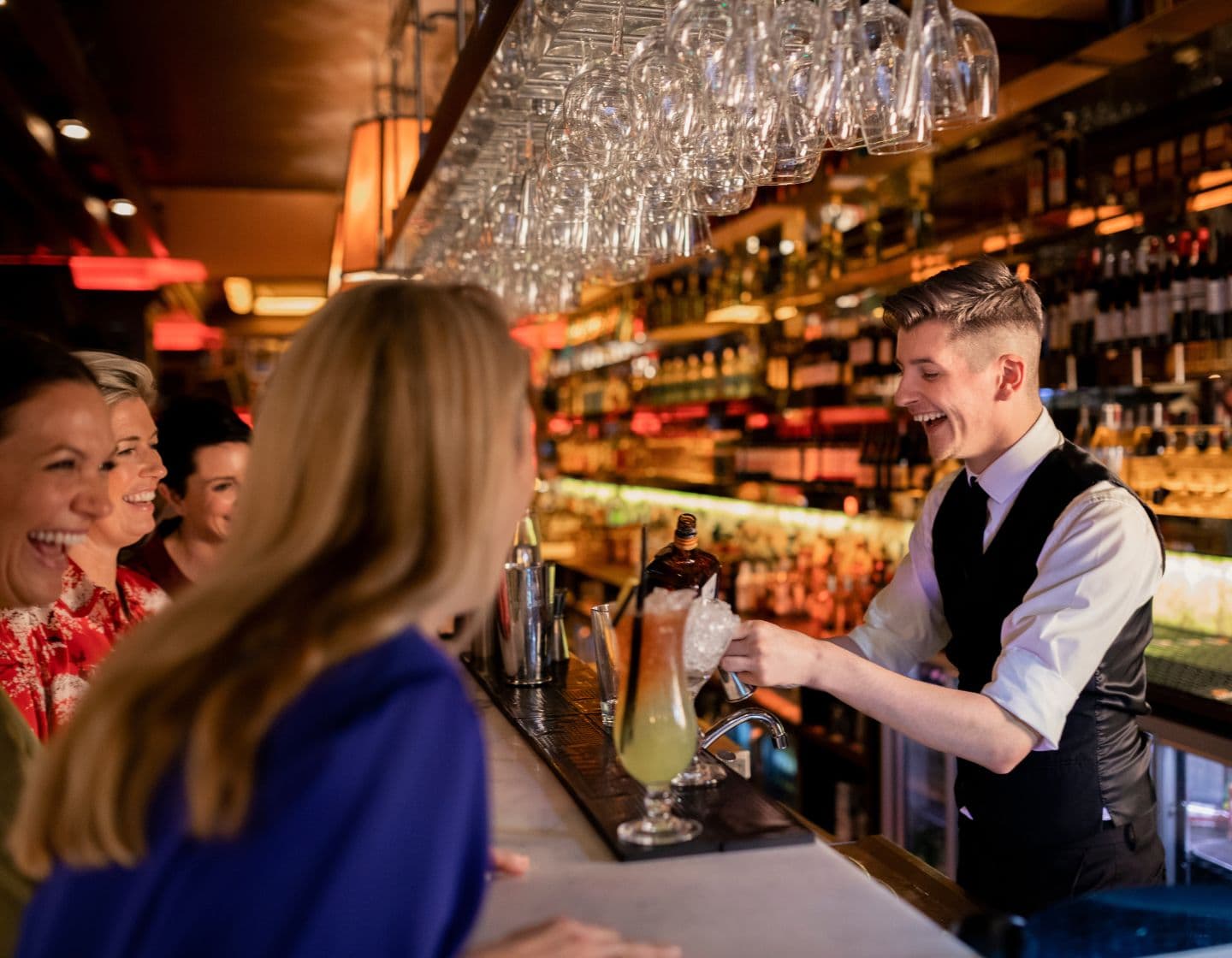 Barkeeper steht hinter der Bar und mixt einen Cocktail, während er mit lächelnden Gästen spricht.