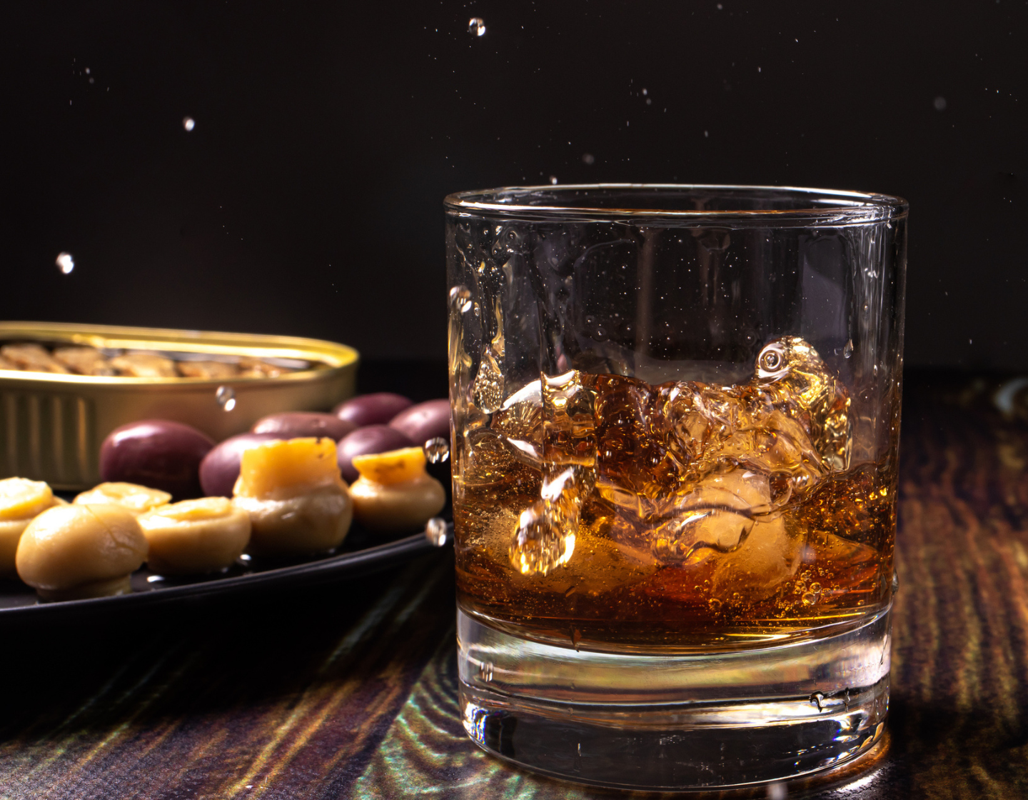 Ein Glas Whisky mit Eis, im Hintergrund ein Teller mit Oliven und Nüssen.