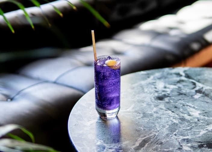 A purple highball cocktail on a round, grey marble table next to a black sofa.