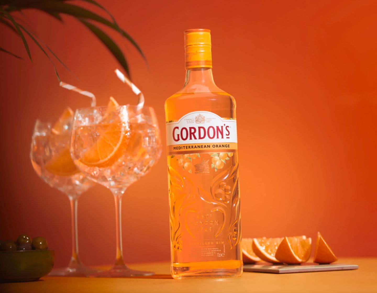 Garrafa de Gordon's Mediterranean gin ao lado de dois copos de G&T em um fundo laranja.