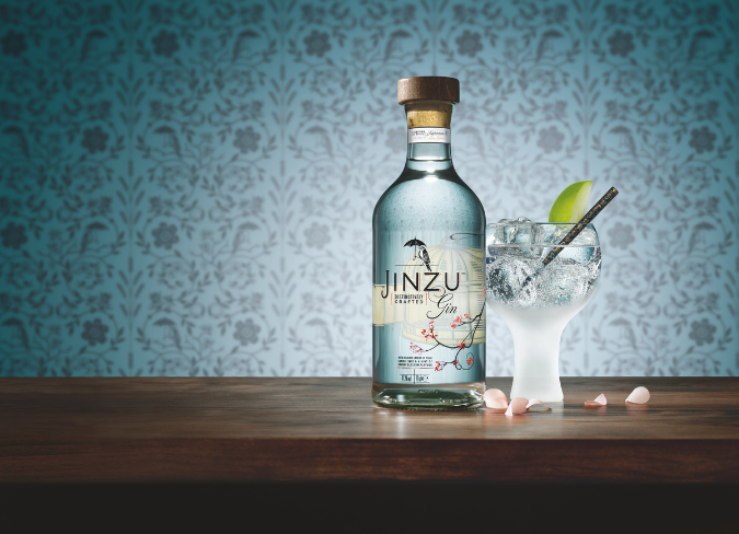 Garrafa de gin Jinzu na mesa ao lado de copo de gin e tônica contra papel de parede floral azul ao fundo 