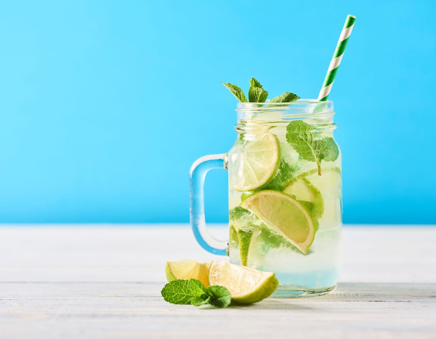 A mojito served with a green and white paper straw against a blue background. 